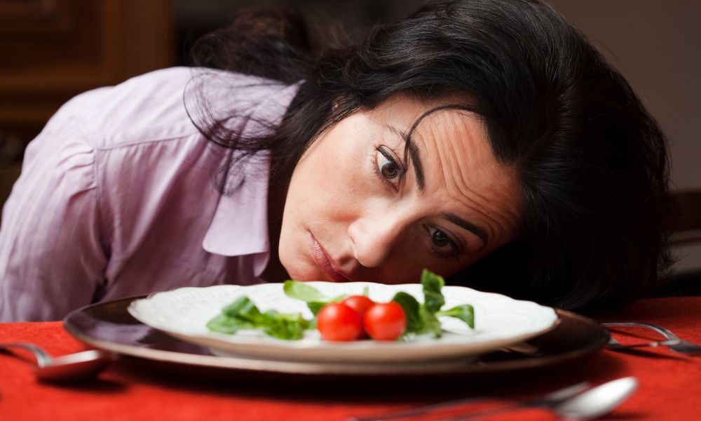 A woman stairs forlonly at a white plate, it contains a few salad leafs and some small tomatos, the plate sits on red table cloth. For blog: risks of fad dieting